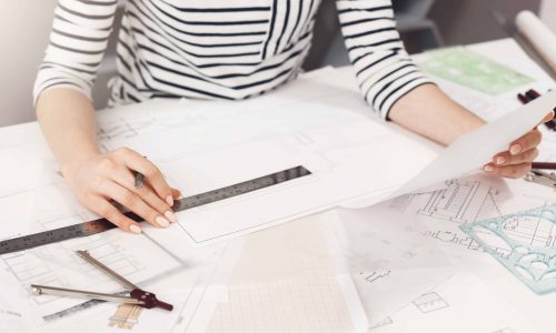 Business concept. Close up detail of young successful architect entrepreneur in striped clothes sitting at white table, looking through work plan, holding pen and ruler in hands, working in new business project in co-working space